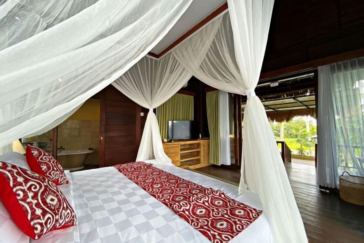 Un dormitorio con cama, dosel blanco, colcha estampada en rojo y blanco, y un televisor sobre un soporte de madera.