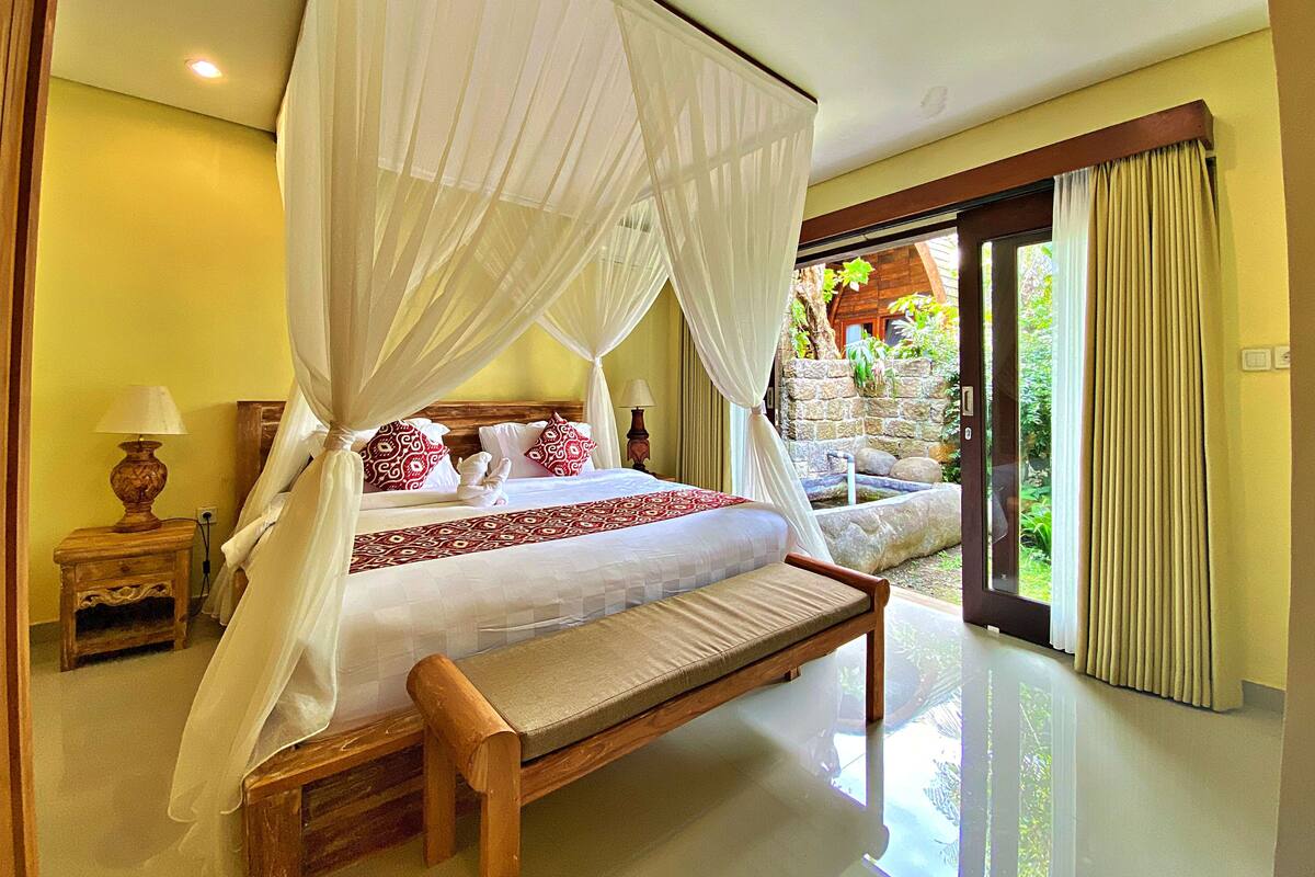 A bedroom with a four-poster bed, a bench, a nightstand, and a view of a garden through a glass door.