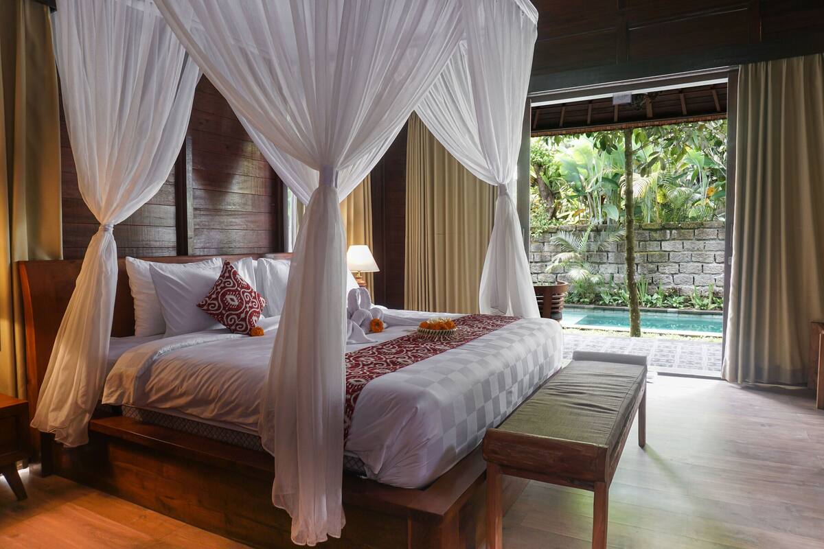 A bedroom with a canopy bed, a bedside table with a lamp, and a view of a pool and greenery outside.