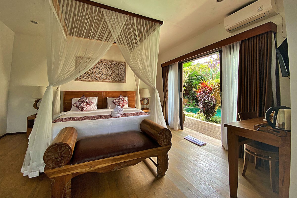 A bedroom with a four-poster bed, a wooden bench, a desk, and a view of a garden through a sliding glass door.