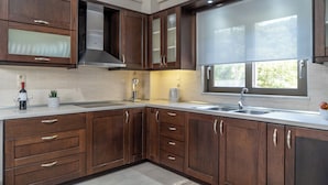 A modern kitchen with dark wooden cabinets, a stainless steel range hood, a large window, and a built-in sink. - Villa Emily (Voukolies, Chania)