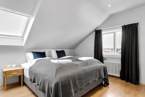 A modern bedroom with a sloped ceiling, a large bed with a gray and black checkered bedspread, white and blue pillows, a wooden nightstand, and a window with black curtains.