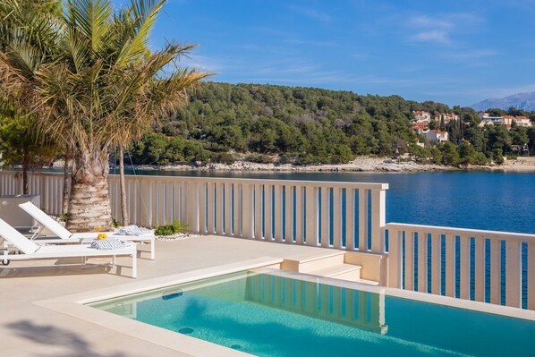 A pool area with lounge chairs, a palm tree, and a view of a coastal landscape with buildings. - More Brac in Selca With 4 Bedrooms and 4 Bathrooms (Selca)