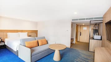Una habitación de hotel con una cama, un sofá gris con cojines anaranjados, una mesa redonda de madera y una alfombra azul.