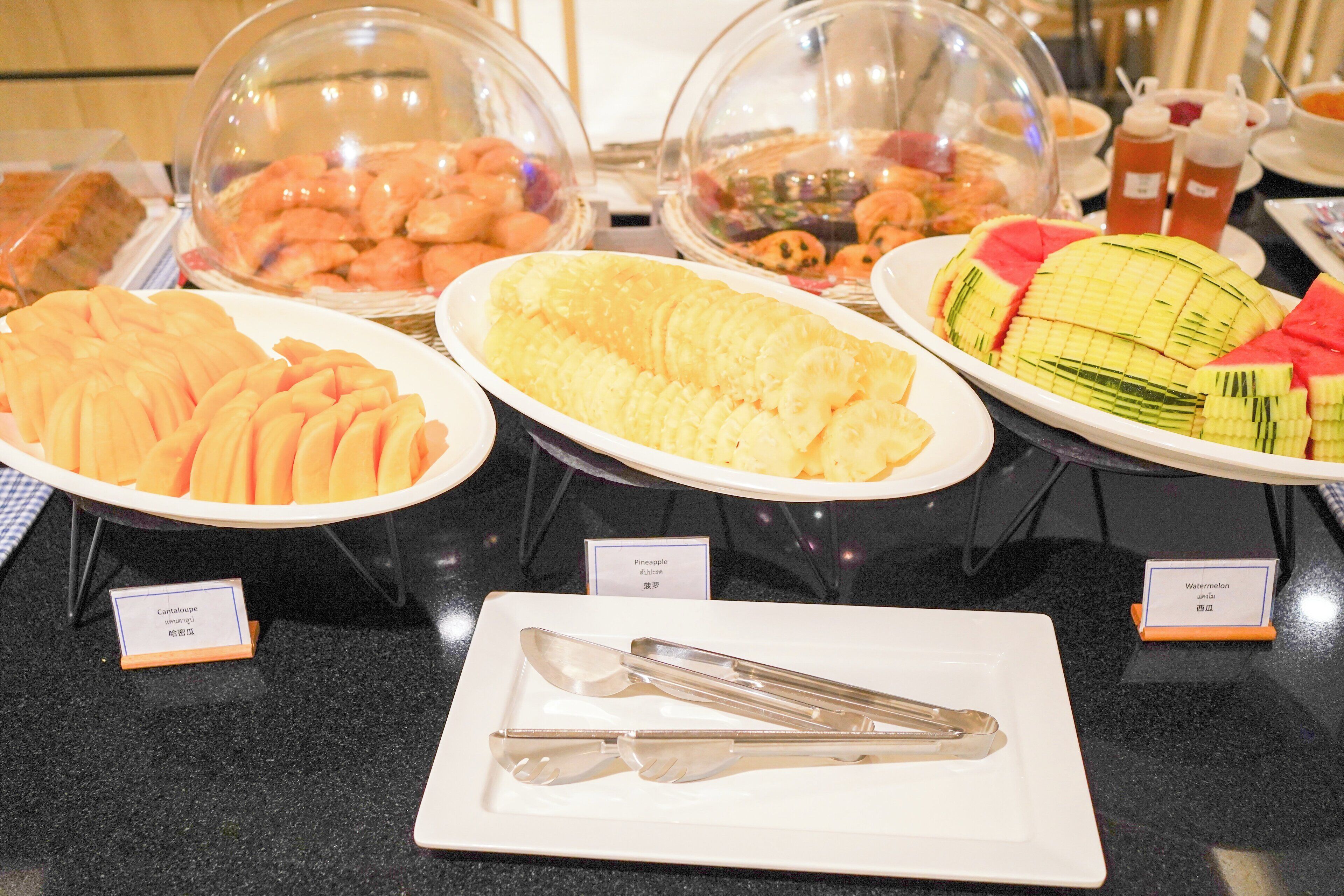 Un buffet proposant une variété de plats présentés sous des cloches de verre, notamment de l’ananas, de la pastèque et d’autres fruits.