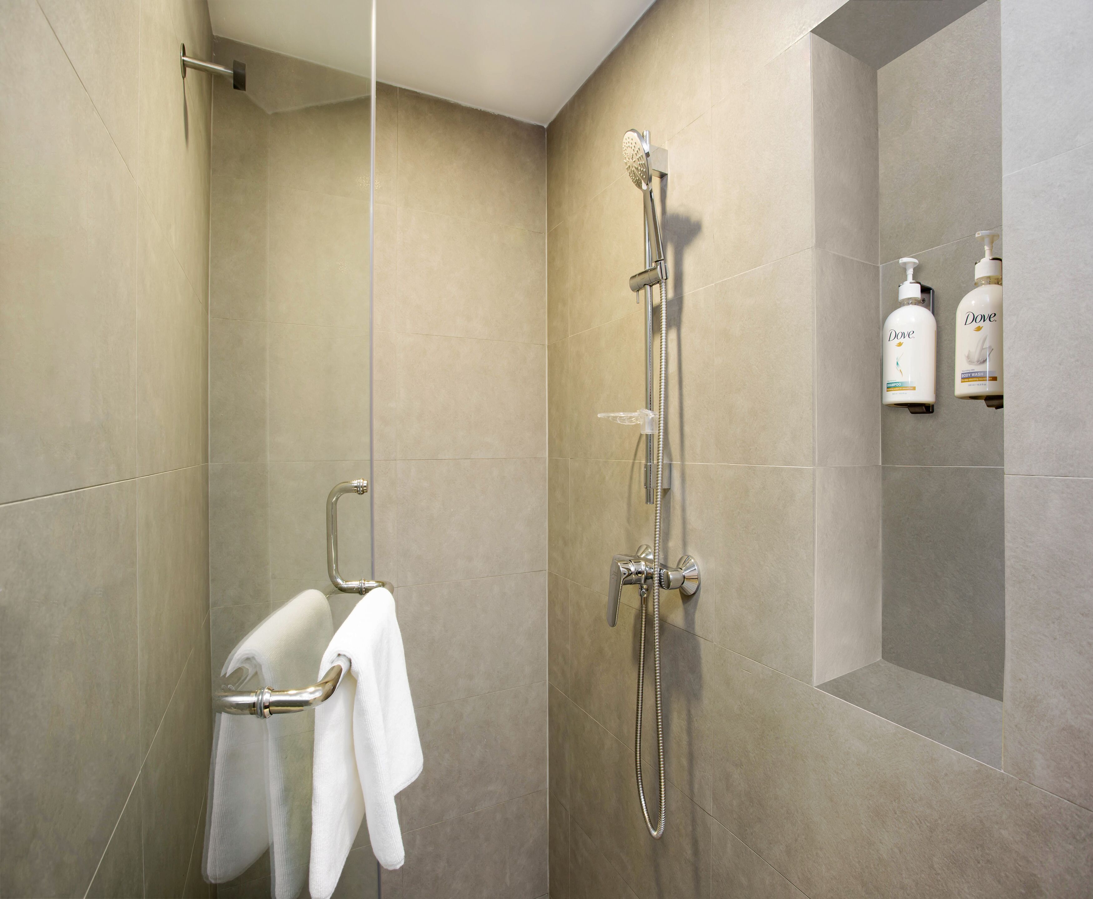 A modern bathroom with beige tiles, a shower with a handheld showerhead, a towel rack with two towels, and Dove hand soap dispensers on the wall.
