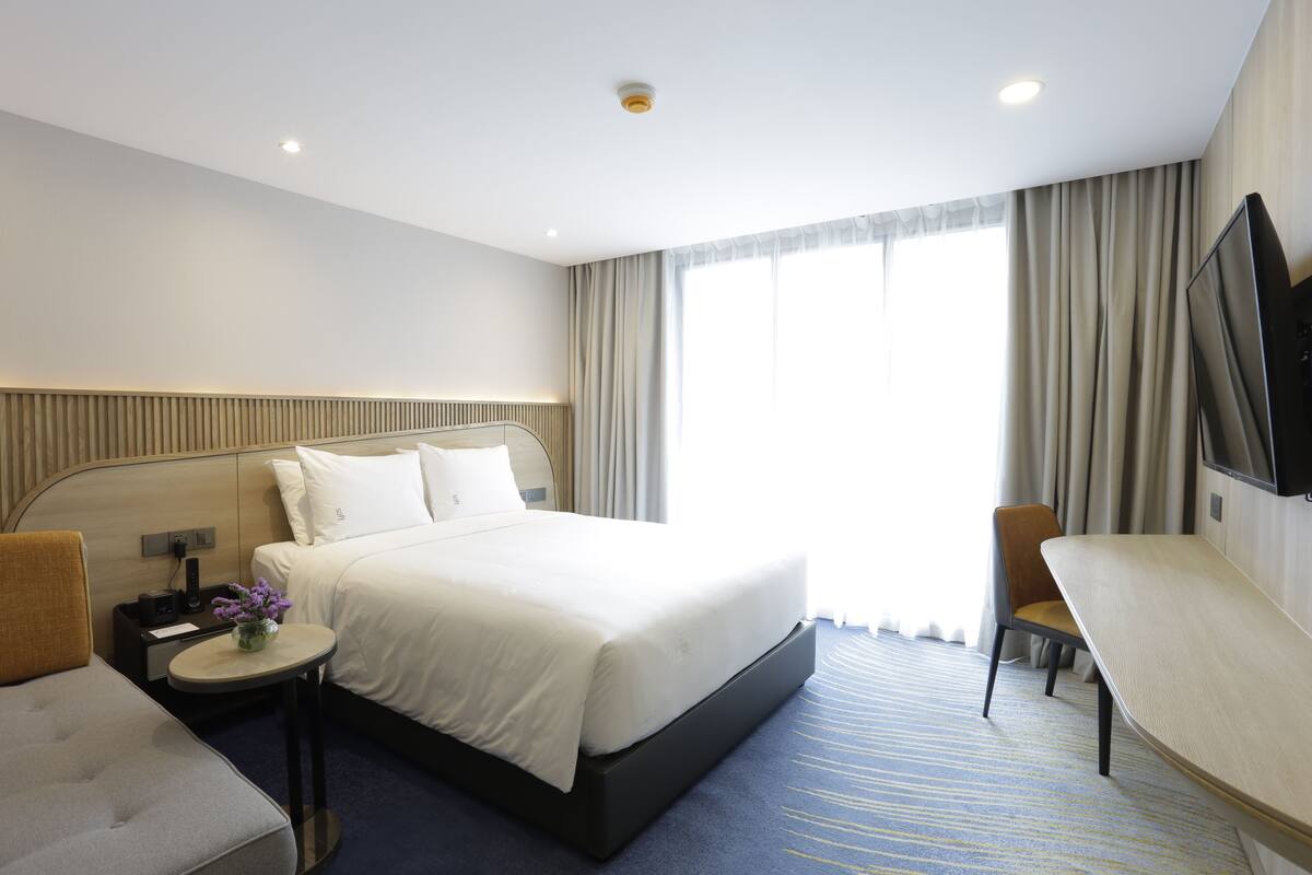Een hotelkamer met een bed, een bureau, een stoel, een kleine tafel met een bloevas en een televisie aan de muur.
