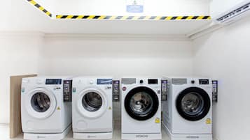 Four white Hitachi washing machines in a row, with a caution sign on the ceiling and an air conditioning unit on the wall.