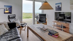 Een moderne woonkamer met een bank, stoelen, een salontafel en een televisie. Er is een groot raam met uitzicht op het strand.