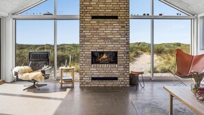 Amplia habitación con una chimenea grande, un sillón de cuero y vistas a las dunas a través de grandes ventanales.