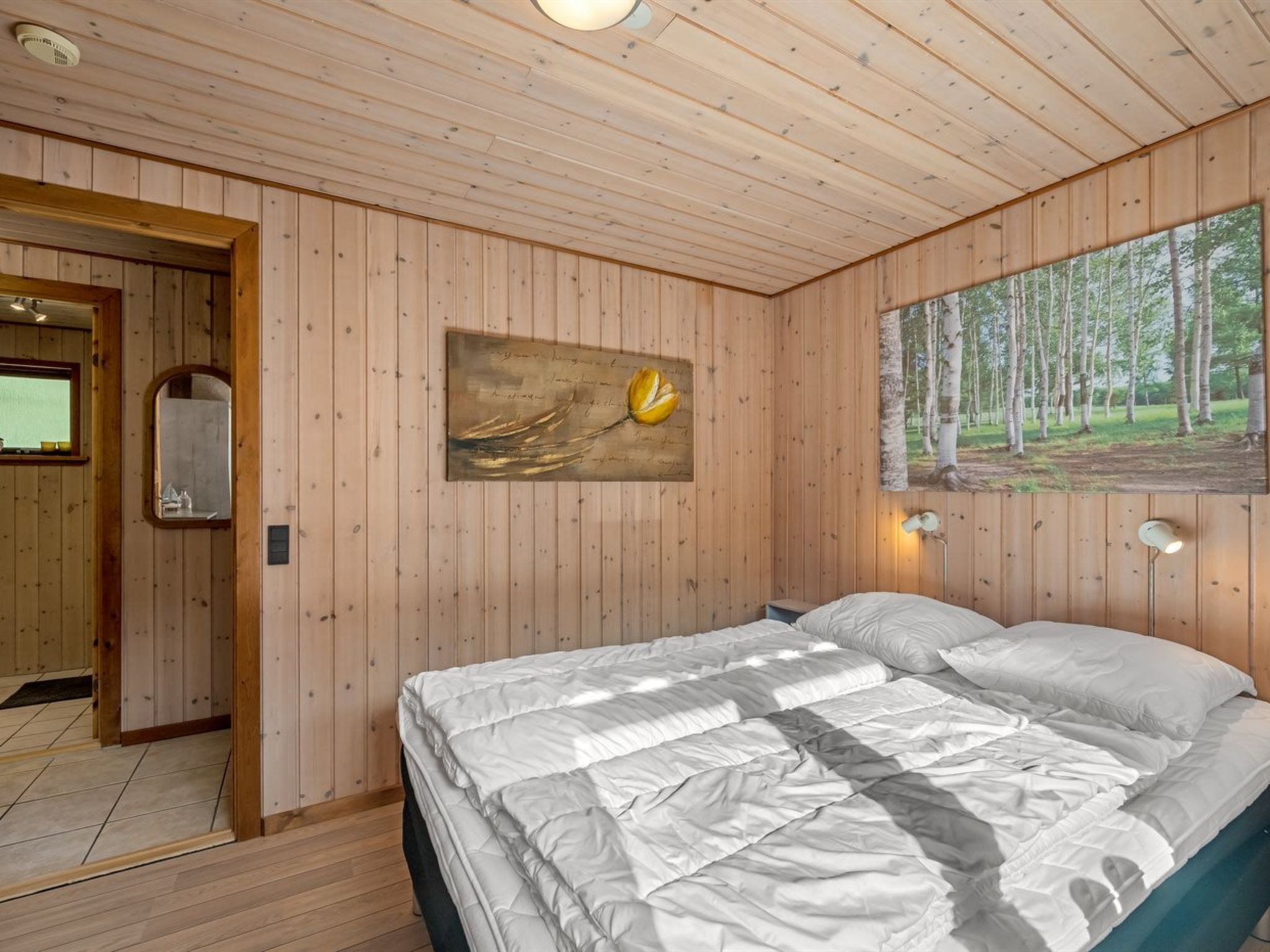 A wooden-paneled room with a bed, a painting of a flower, and a forest view.