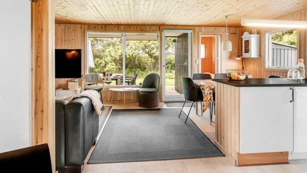 A modern cabin interior with a wooden ceiling, a cozy seating area with a sofa and chairs, a kitchen with a bar counter, and large sliding glass doors leading to an outdoor patio. - "Bistra" - 400m from the sea by Interhome (Løkken)