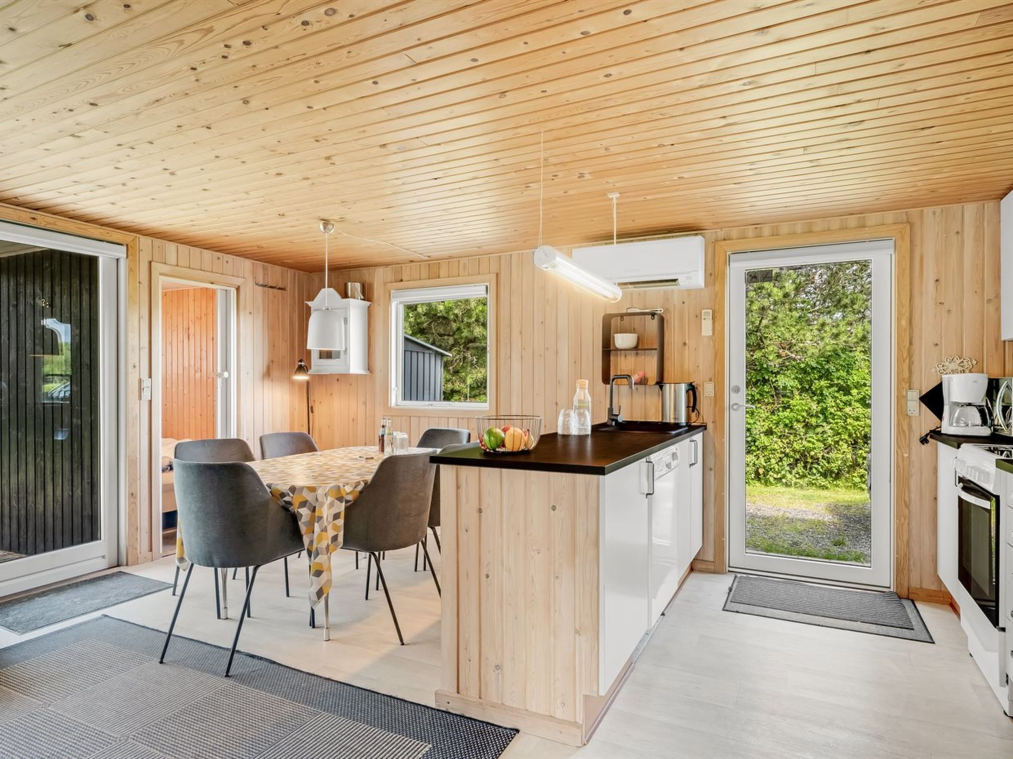 Una cocina moderna con una isla central, comedor con sillas, paredes de madera y ventanas grandes.