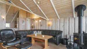 A cozy cabin interior with a wooden ceiling, a black leather sofa, a wooden table, and a wood-burning stove.