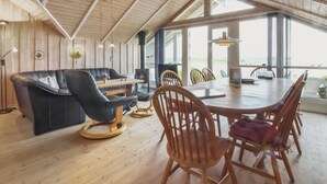A spacious living area with a wooden ceiling, a black leather sofa, wooden chairs, a dining table, and large windows offering a view.