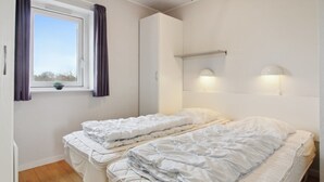 A bedroom with two beds, a window with curtains, a bedside table, and a wall-mounted light fixture.