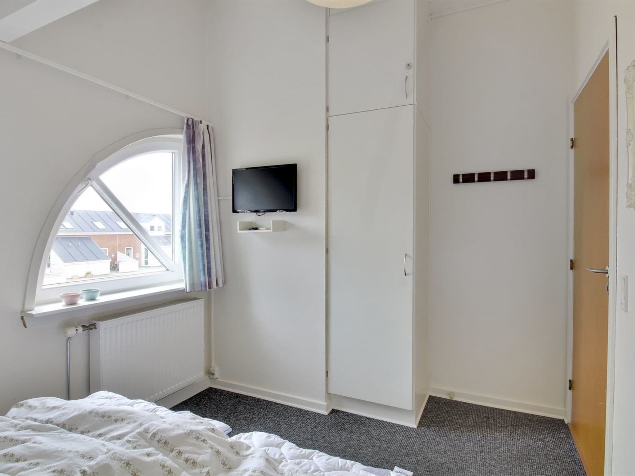 Un dormitorio con una cama, televisor montado en la pared, armario y ventana con cortinas.