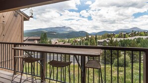 Un balcone con sedie in vimini e un tavolo di legno, con vista su un paesaggio montuoso con edifici e alberi.