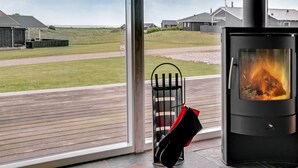 A modern interior with a wood-burning stove, large windows, and a view of a grassy field and houses.