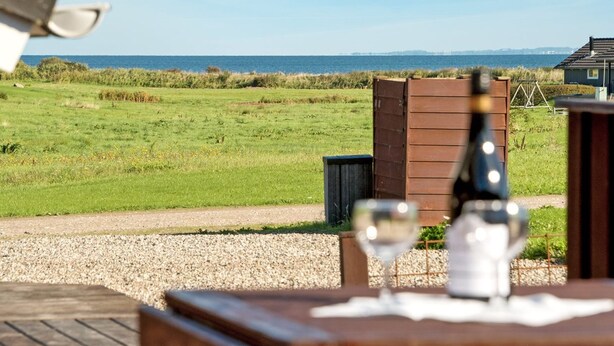 A table with two wine glasses and a bottle, set against a backdrop of a grassy field and a wooden structure, with a clear view of the sea and distant buildings.