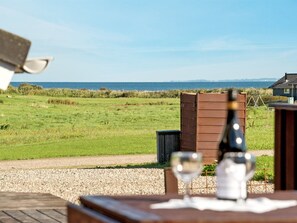A table with two wine glasses and a bottle, set against a backdrop of a grassy field and a wooden structure, with a clear view of the sea and distant buildings. - "Alexis" - 250m from the sea by Interhome (Nordborg)