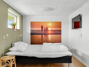 A bedroom with a bed, a window with a plant, a wall-mounted TV, and a sunset painting.