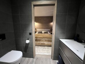 A modern bathroom with a white toilet, a large bathtub, and a wooden door leading to a bedroom with a bed and a nightstand. - Stylish Penthouse in Tignes Lavachet, 4 Bedrooms, WiFi, 8 People (Tignes)