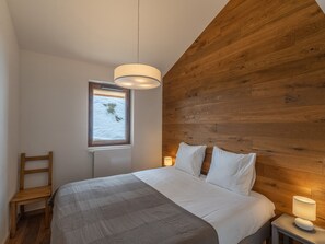 Ein Schlafzimmer mit hölzernem Kopfteil, einem Bett mit weißer Bettwäsche, einem Nachttisch mit Lampe, einem Fenster mit Blick ins Grüne und einem Holzstuhl.