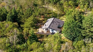 A house surrounded by dense forest, with a sloped roof and visible outdoor seating area.