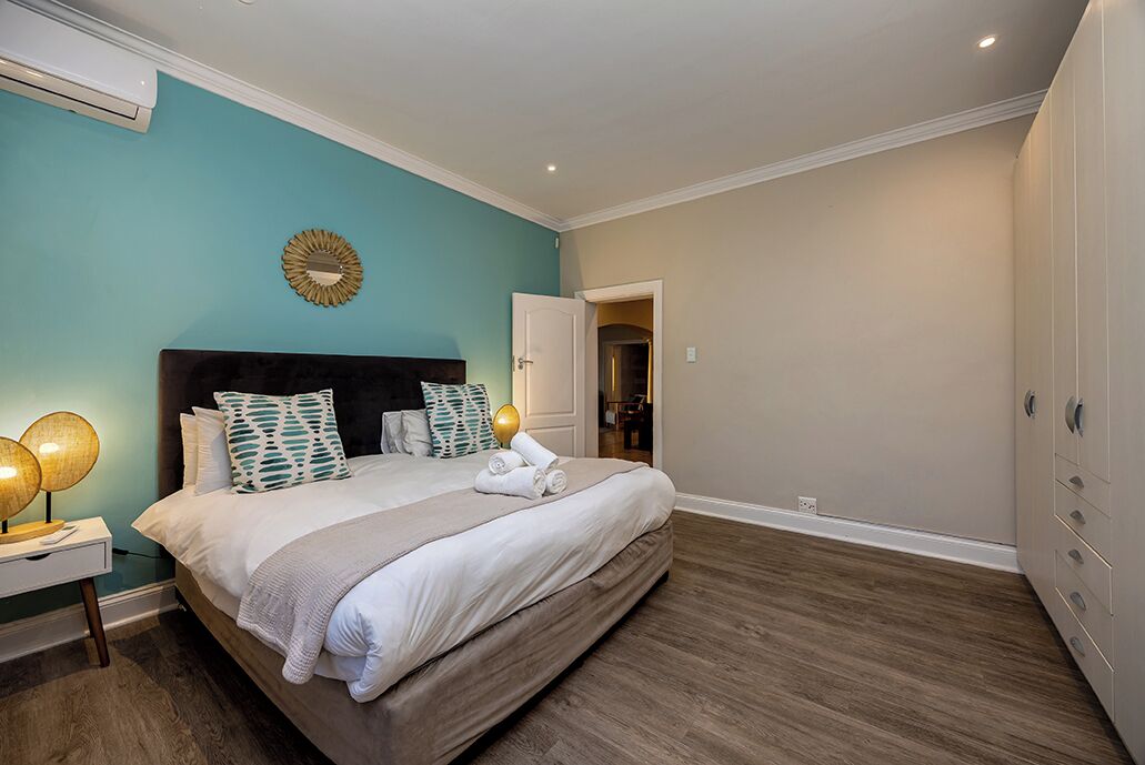 A bedroom with a large bed, a nightstand with lamps, a wall clock, and a door leading to another room.