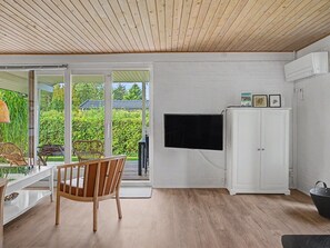 Een kamer met een houten vloer, een witte kast, een aan de muur bevestigde televisie en een glazen schuifdeur die leidt naar een buitenruimte met groen.