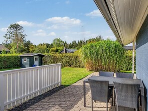 A patio area with a table and chairs, a white fence, and a garden with a hedge and a small shed.