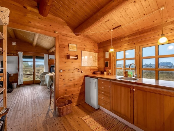 A wooden cabin interior with a kitchen, dining area, and a view through the windows.