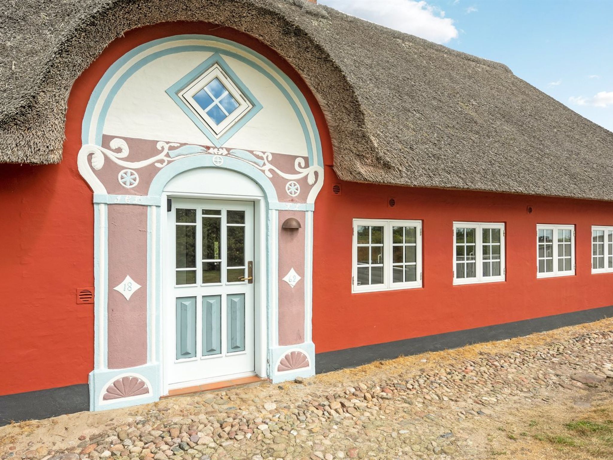 Una casa tradicional con techo de paja, fachada roja y puerta blanca, con detalles decorativos y ventanas numeradas.