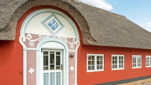 Une maison traditionnelle à toit de chaume, avec une façade rouge et une porte blanche, agrémentée d’éléments décoratifs et de fenêtres numérotées.