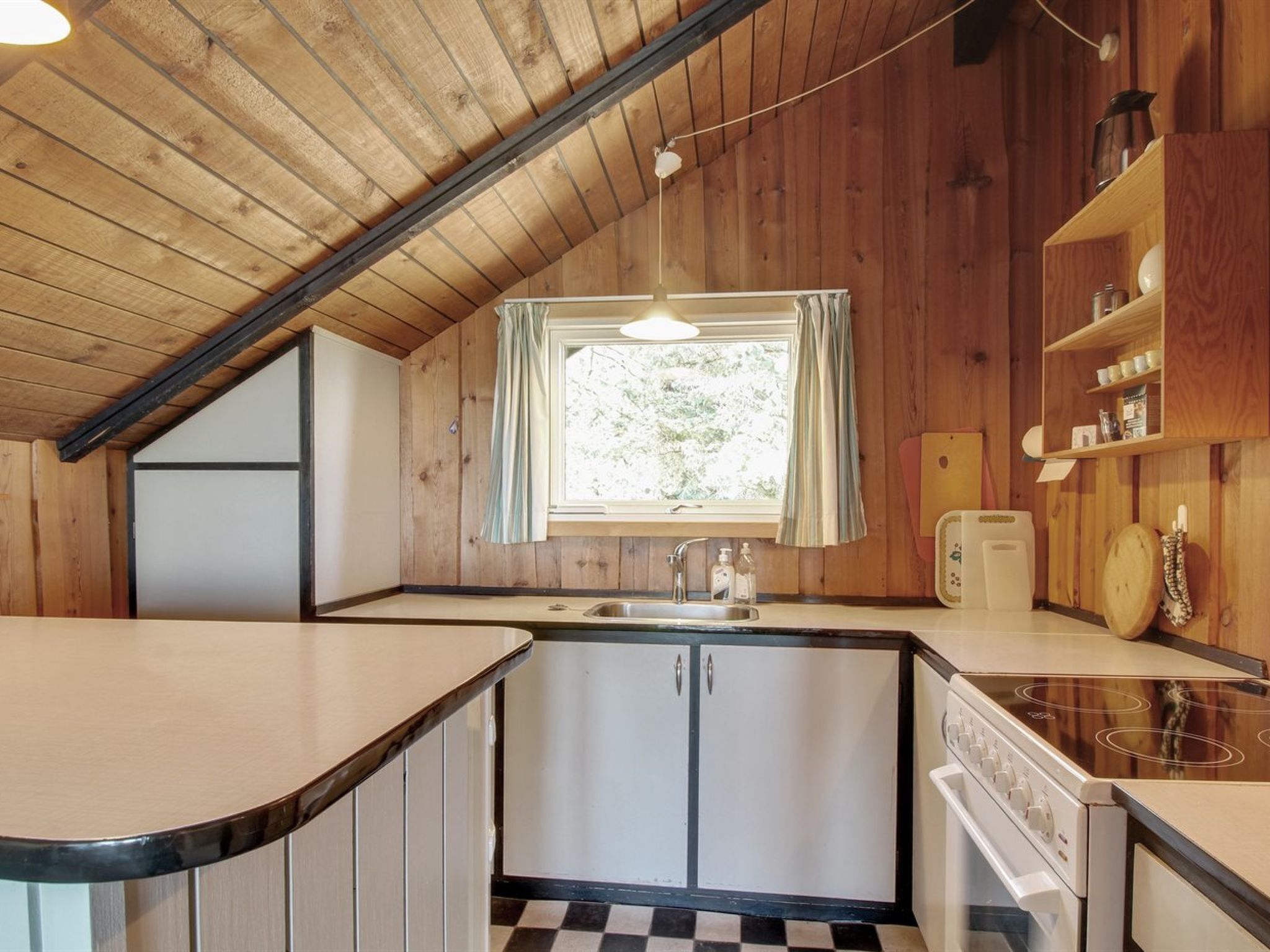 A compact kitchen with a central island, a window with curtains, wooden walls, and a checkered floor.