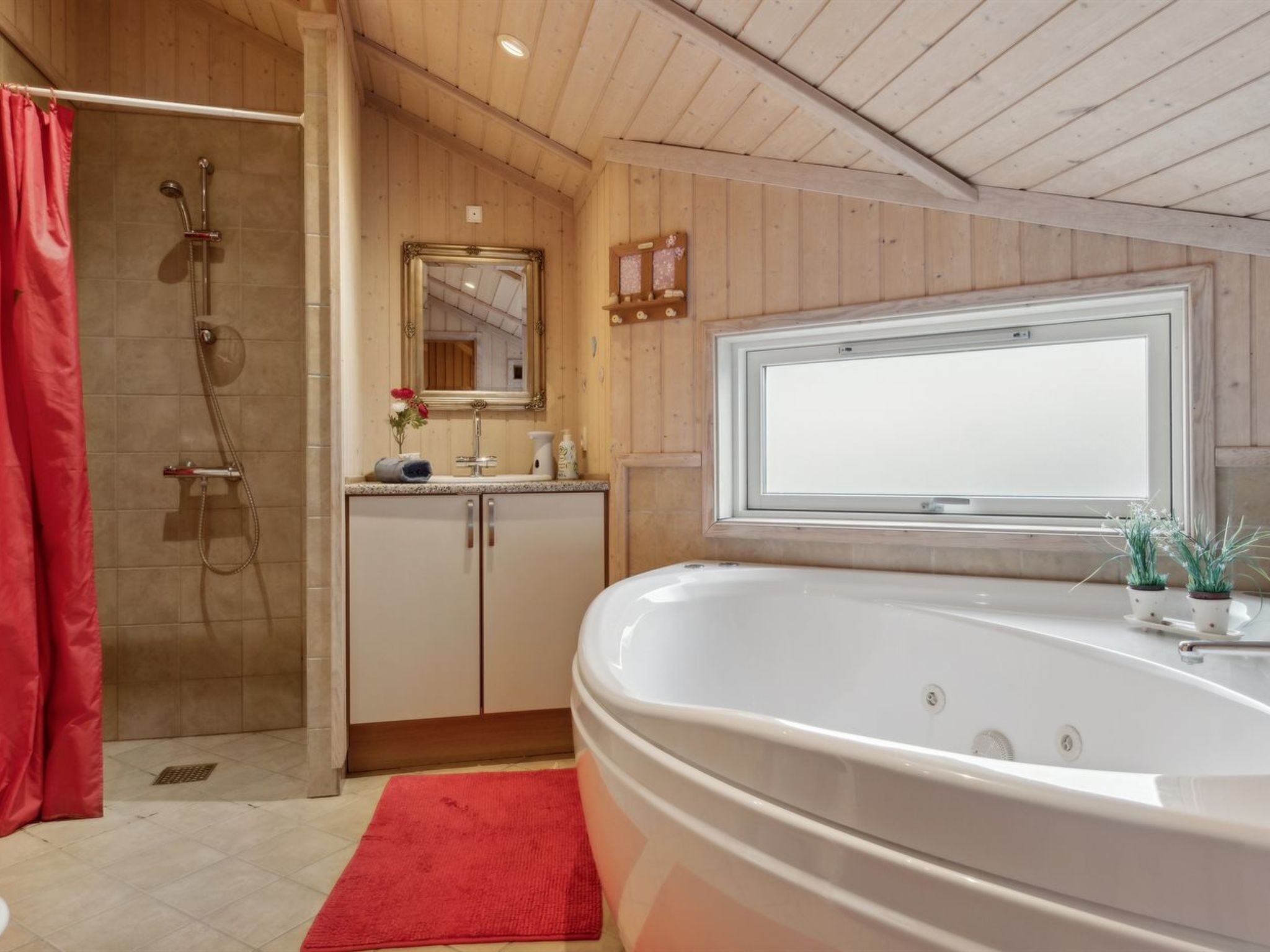 A bathroom with a large bathtub, a shower, wooden walls, a window, and a red bath mat.