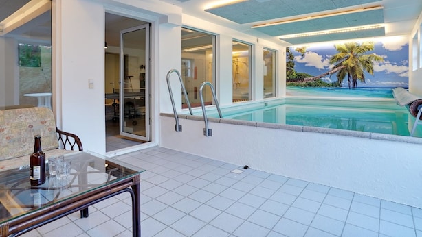 Ein moderner Innenpoolbereich mit Glaswand, ein Pool mit Blick auf einen tropischen Strand und eine Sitzecke mit Tisch und Stühlen.