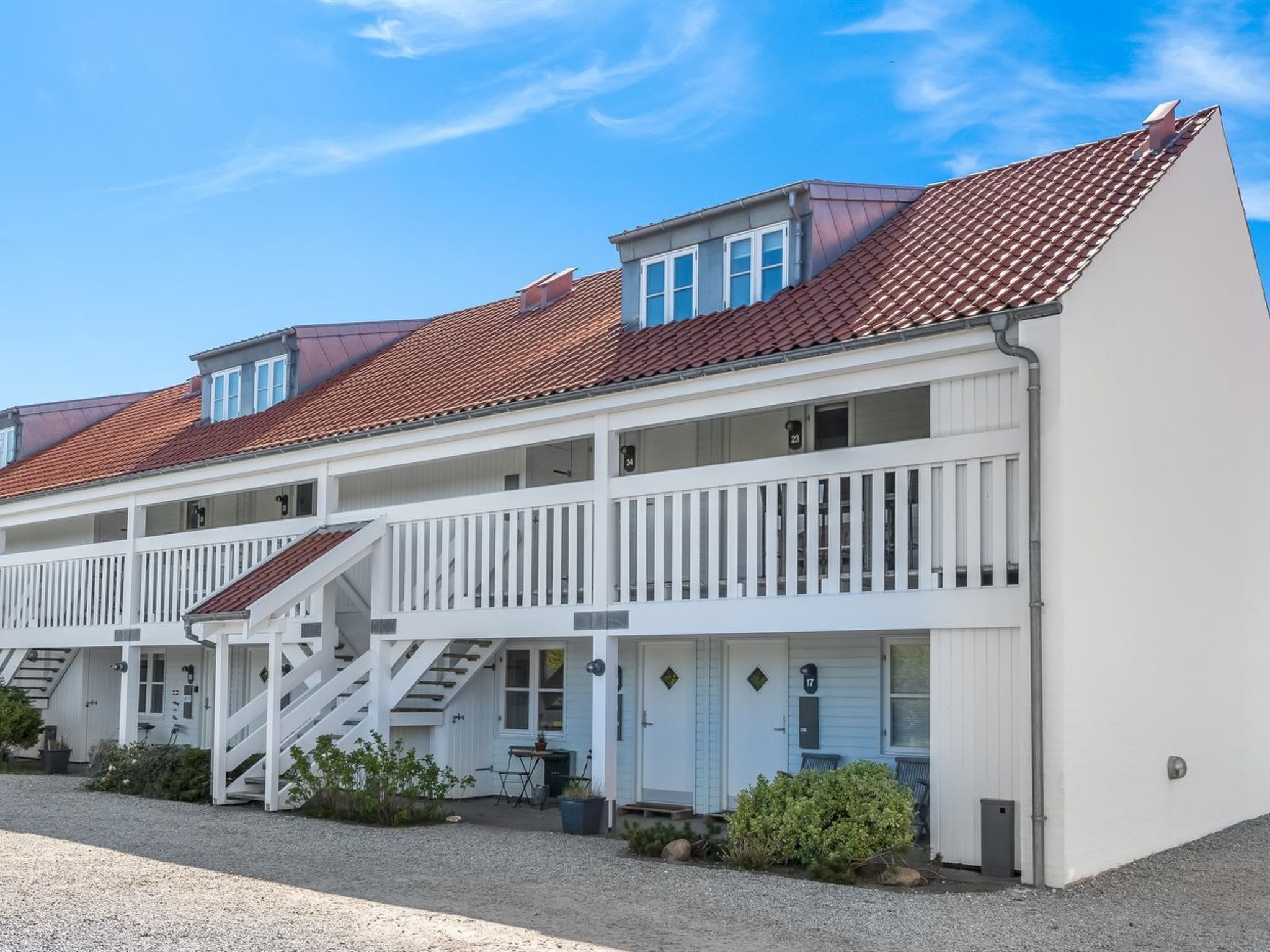 A row of white, two-story buildings with red-tiled roofs, featuring balconies and a gravel driveway.