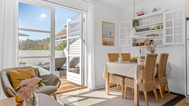Un espace de vie et de salle à manger lumineux et décloisonné, avec une porte-fenêtre coulissante donnant sur un balcon, des meubles en osier et un meuble blanc contenant de l’ustensile de cuisine.