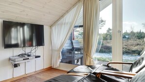 Een kamer met een flatscreen televisie, houten plafond, een bank en uitzicht op een natuurlandschap door een glazen deur.