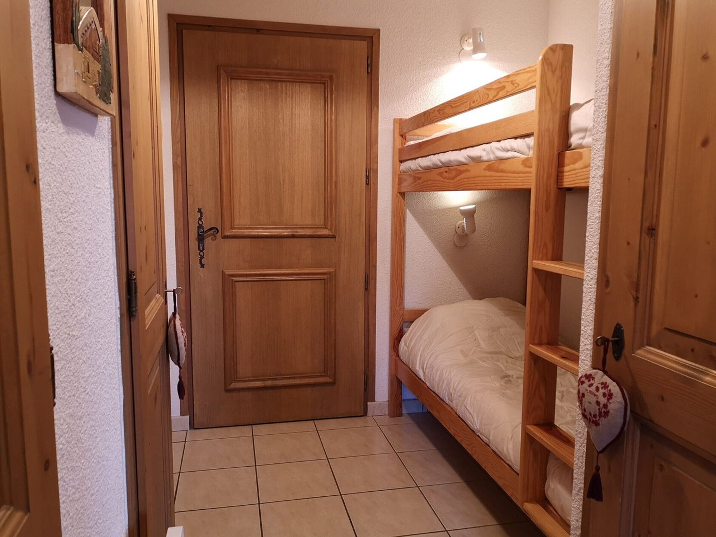 A wooden bunk bed with a headboard and a small shelf, a door with a brass handle, and a wall-mounted light fixture.