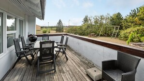 Een overdekt terras met een houten dek, een tafel met stoelen en een rotan fauteuil.