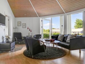 Ein modernes Wohnzimmer mit Holzboden, großen Fenstern, einem schwarzen Ledersofa, einem Esstisch und einem Fernseher.