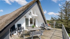 A thatched-roof house with a wooden deck, outdoor seating, and a view of the surrounding landscape.