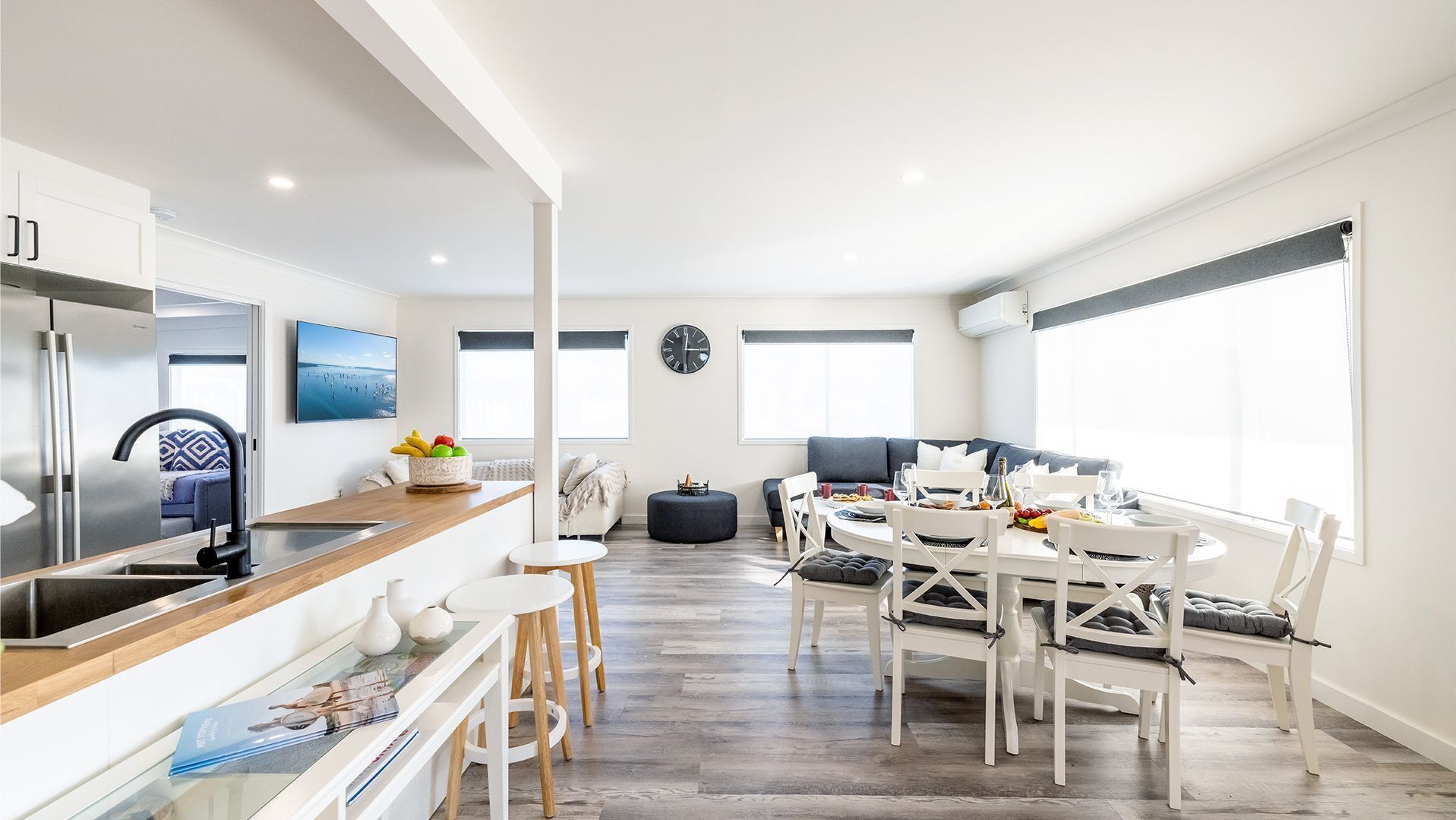 Una cocina moderna con horno empotrado, un comedor con mesa redonda y sillas blancas, y una sala de estar con sofá y un ventanal grande.