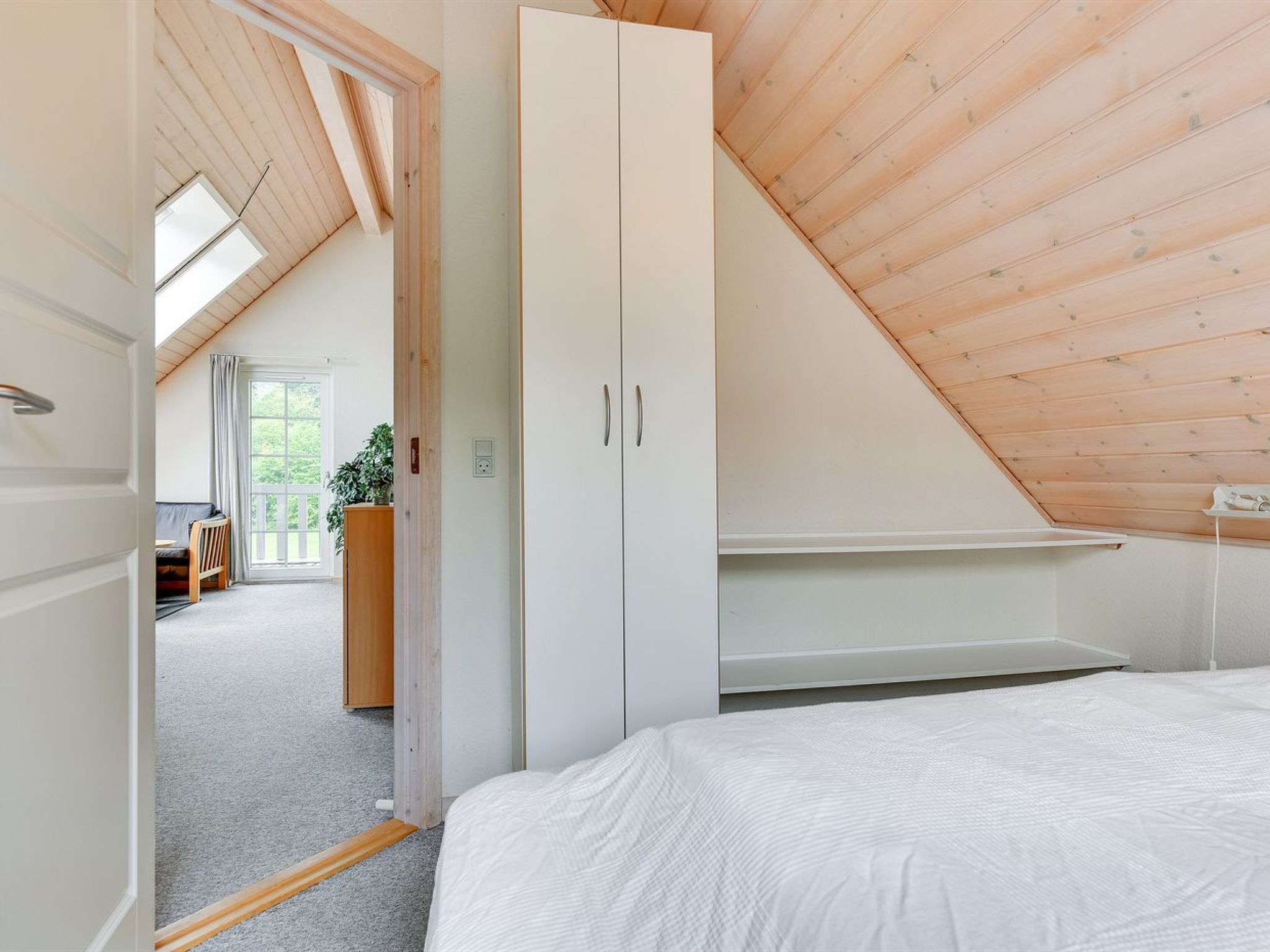 Een slaapkamer met schuine wanden, een bed, een garderobe en uitzicht op een buitenruimte met een plant en een bank.