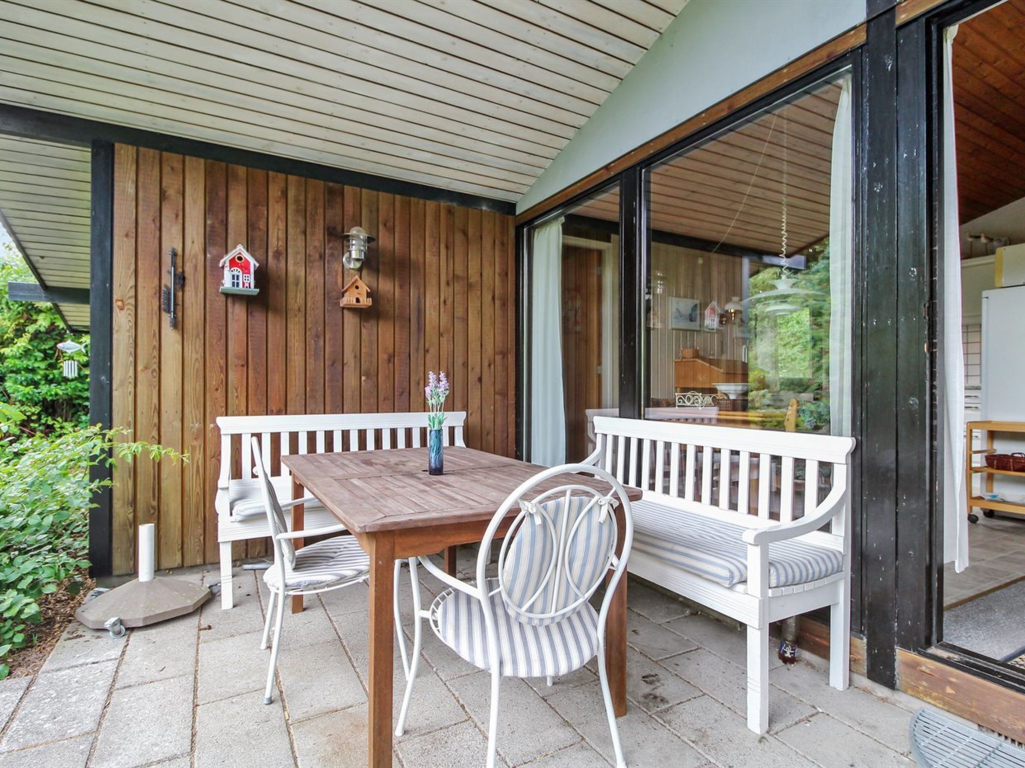 An outdoor patio with a wooden table and white chairs, a white bench, and a sliding glass door leading to a kitchen area.