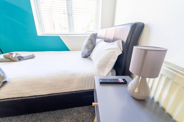 A neatly made bed with pillows, a remote control, and a lamp on a bedside table in a room with a window and a teal accent wall.