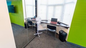 Un petit bureau avec un bureau, deux chaises, un ordinateur portable, un ventilateur et une fenêtre avec des stores.